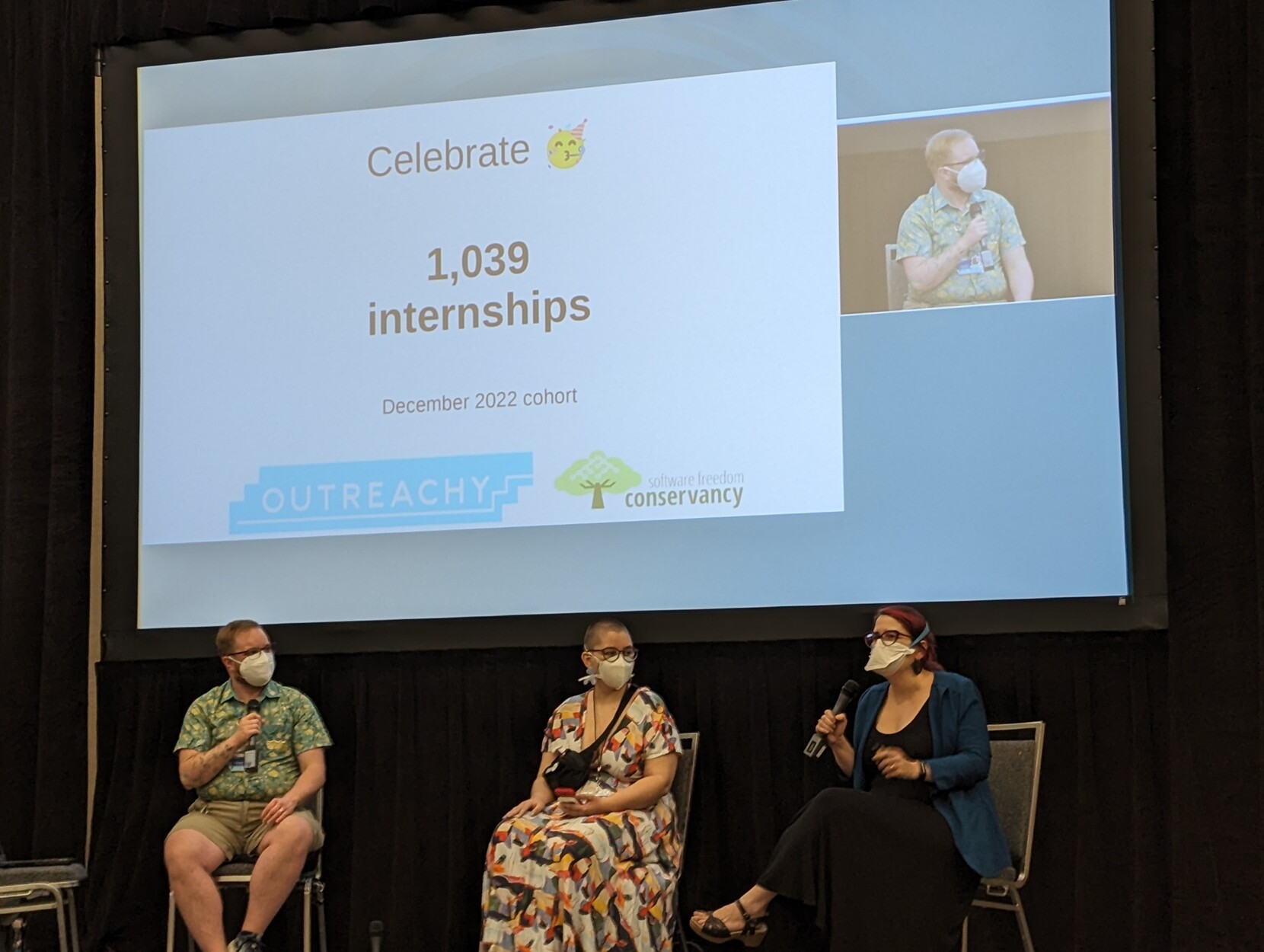 A photo of Sage, Anna, and Karen on stage sitting in front of a big slide noting Outreachy has provided internships for 1,039 people!